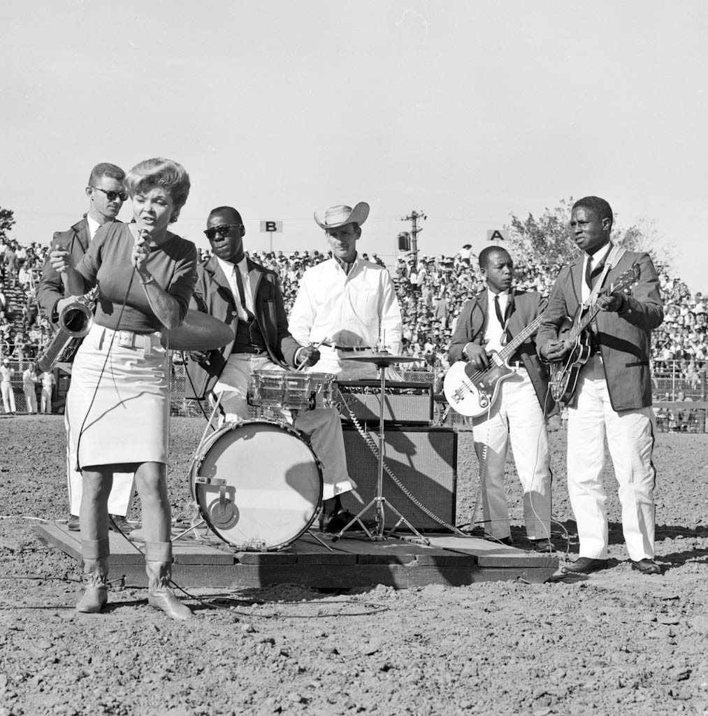 A black-and-white photo showing a White woman with a bouffant hairstyle singing into a microphone on the left side of the frame on the dirt grounds of a rodeo, while in the background are Black and White incarcerated men, wearing dark colored jackets, black ties and billowing white pants playing guitars and a drum. The drummer is wearing sunglasses, as a correctional officer stands guard behind him dressed in white and a cowboy hat with the brim upturned on both sides. In the distance, crowds sit in the stands.
