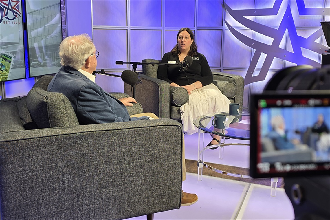 OK Carter and Erica Rousseau seated with microphones in front of them for podcast episode with camera recording in the foreground.