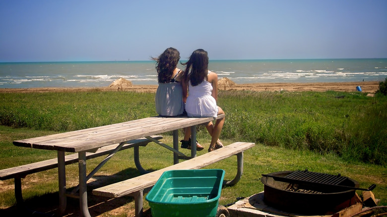 Two girls sitting on a picnic table next to a grill looking out at Quintana Beach with rolling waves and sand.