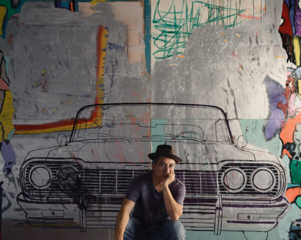A photograph of artist Cande Aguilar squatting and posing in front of a large painting of a car.