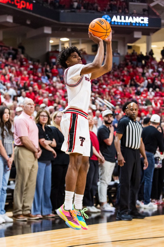 A basketball player in white uniform with red and black stripes, Christian Anderson, jumps to shoot a three-pointer in an arena packed with spectators.