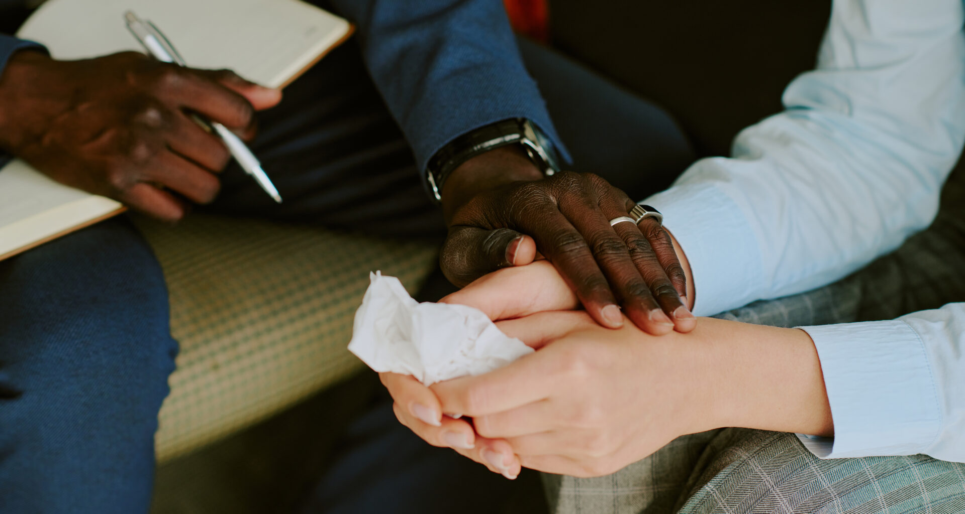 tight photo of a black man's and white woman's hands. The man holds a notepad and pen in one hand and holds the woman's hands with the other. She holds a facial tissue.