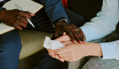 tight photo of a black man's and white woman's hands. The man holds a notepad and pen in one hand and holds the woman's hands with the other. She holds a facial tissue.