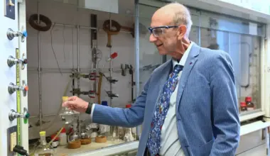 An older male scientist wearing a pinstripe blazer and zany tie poses holding a flask in a scientific lab