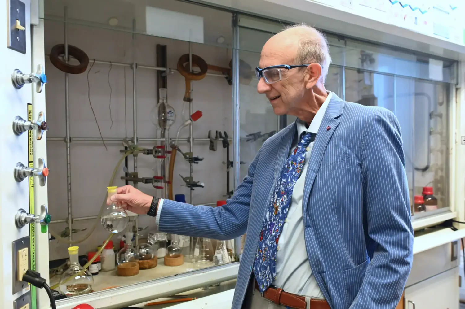 An older male scientist wearing a pinstripe blazer and zany tie poses holding a flask in a scientific lab