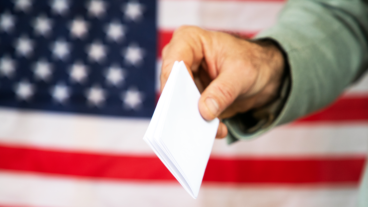 A picture of a hand putting a ballot into a ballot box with the American flag in the background.