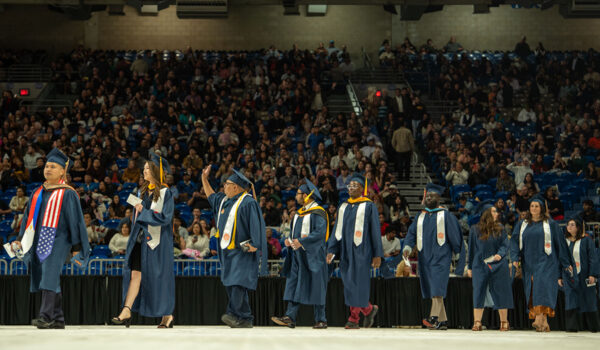 A line of graduates walk toward the stage at the Alamodome during Commencement while a large crowd watches
