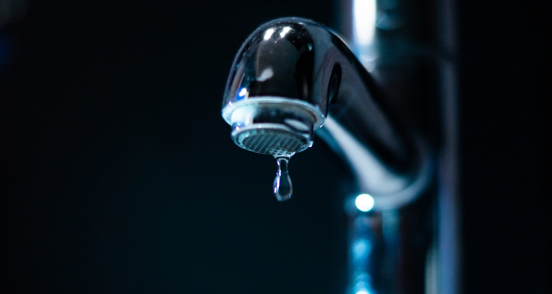 A close-up of a shiny faucet with a single droplet of water hanging from the tip against a dark background.