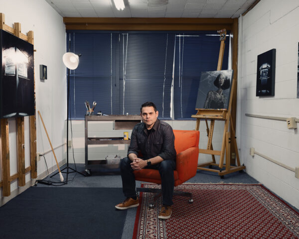 A photograph of artist Joe Peña in his studio/office with artworks behind him.