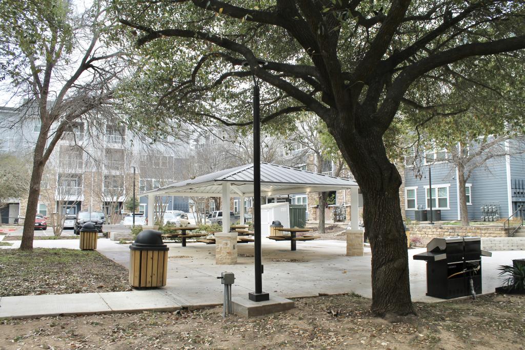 The picture to the right shows a covered picnic pavilion in the background and a large tree, a barbecue grill and trash cans in the foreground. 