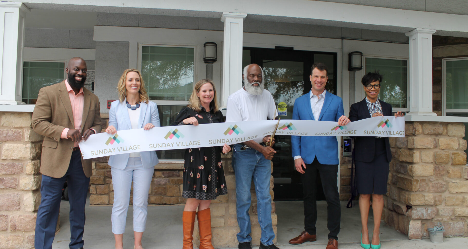 Image shows five individuals holding a long white ribbon with the repeating words Sunday Village. A man in the center holds large gold scissors and is cutting the ribbon.