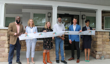 Image shows five individuals holding a long white ribbon with the repeating words Sunday Village. A man in the center holds large gold scissors and is cutting the ribbon.