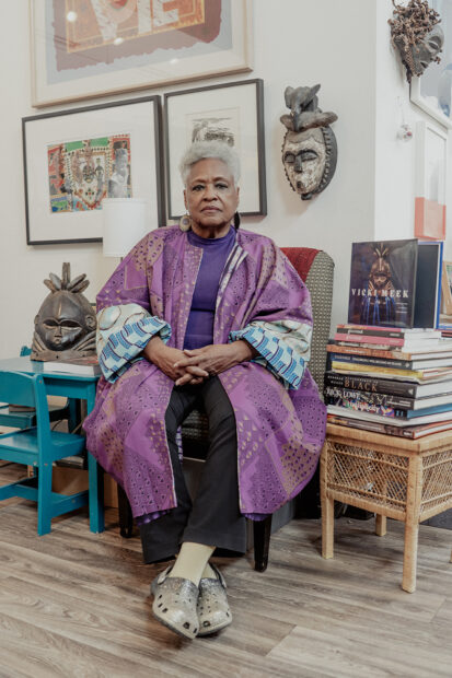 A photograph of artist and cultural worker Vicki Meek sitting in a chair, with a stack of books near her and artworks hung on the wall behind her.