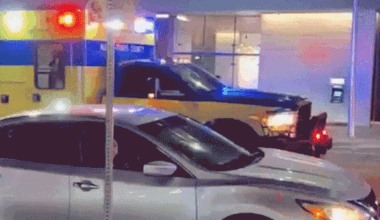 A Waymo autonomous car pauses near an emergency vehicle with flashing lights outside a downtown Austin garage at night.