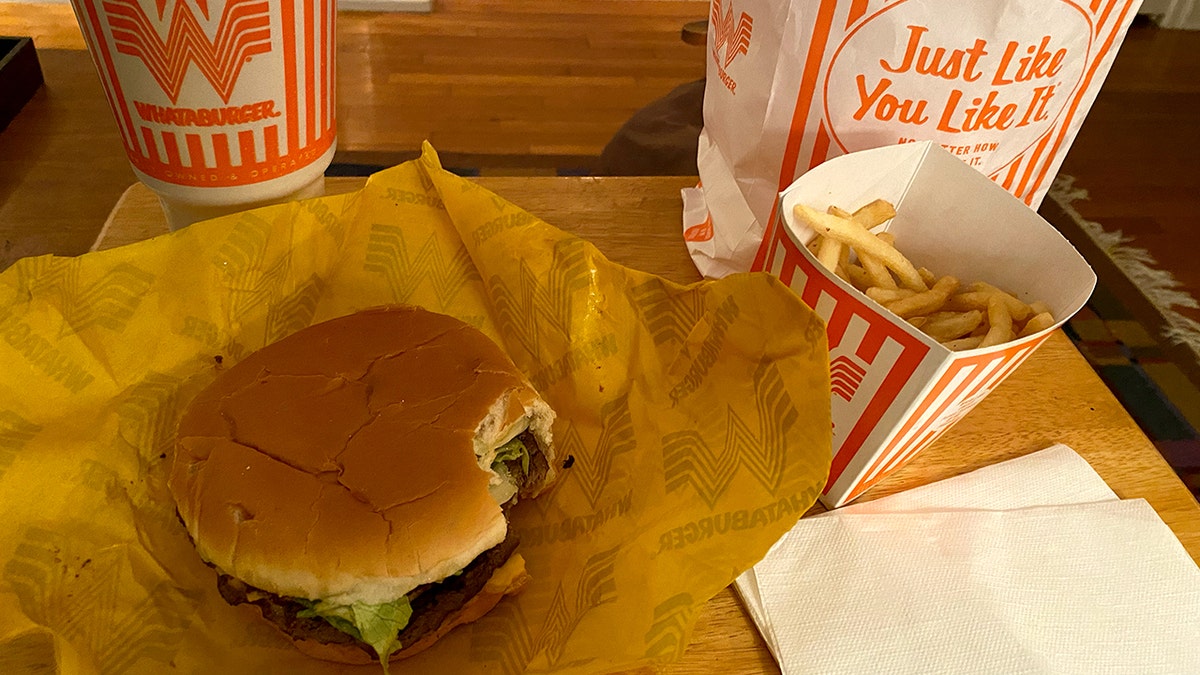 A Whataburger meal with fries and a drink are shown on a TV tray. The burger has a bite in it.
