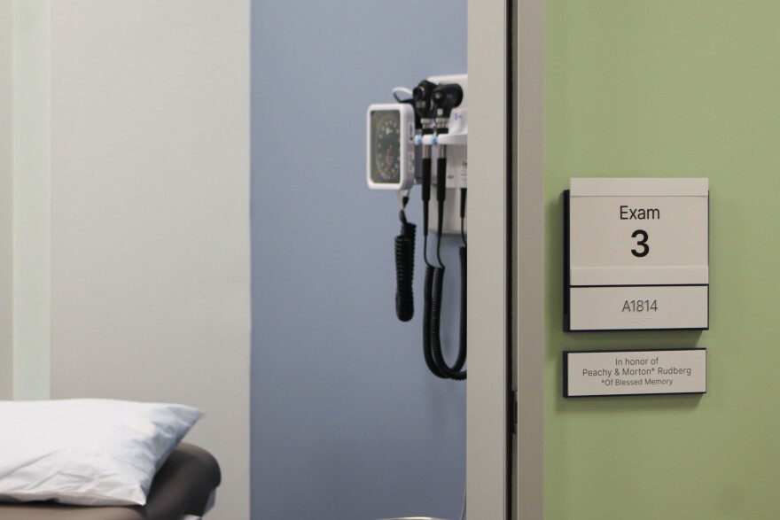 An exam room in the JFS Dallas Parkside Health Center. A pillow rests on a grey exam table and there are medical instruments affixed to a blue wall. A sign on the wall outside the exam room says "Exam 3."