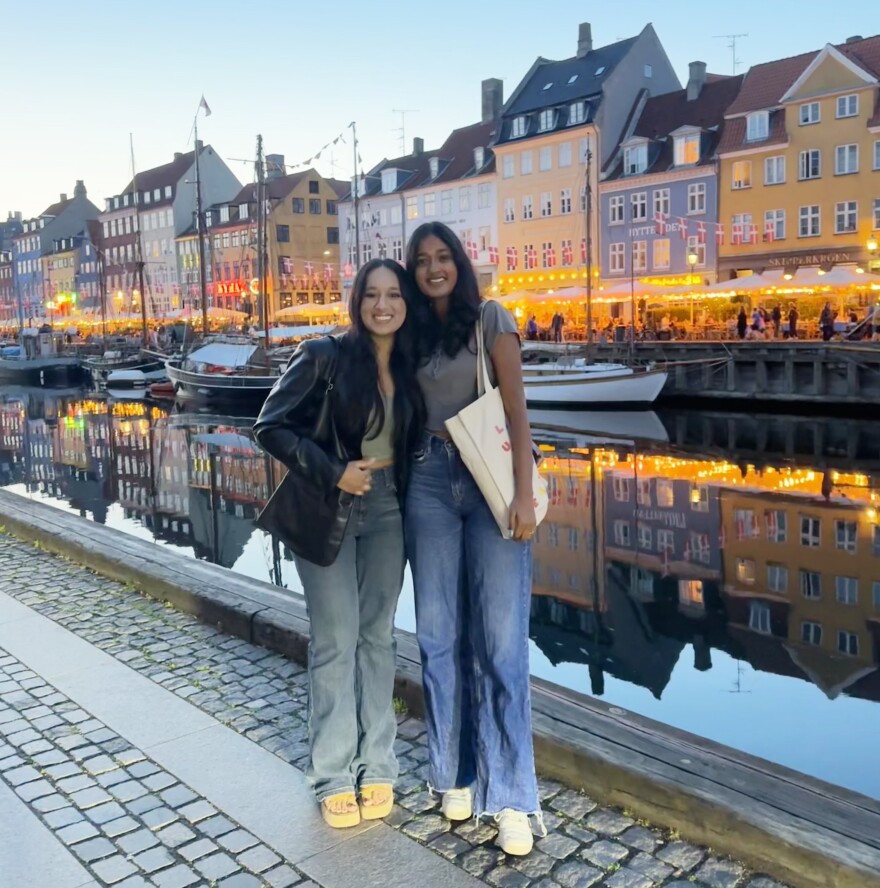 Two women stand on a street with a canal and colorful houses behind them. 