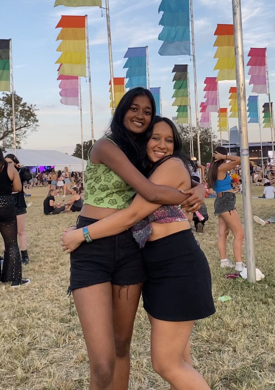 Two women hug in front of colorful flags at a music festival. 