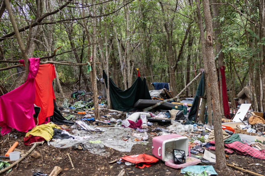 A wooded area is show with turned over tents, coolers and other belongings at a homeless encampment. 