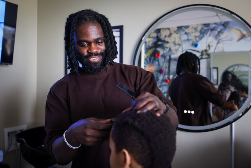 Henry "Tribes" Amoloja, a Black man in a brown shirt, cuts Isaiah Johnson’s, 10, hair. Johnson is visible from behind. There is a mirror behind Henry.