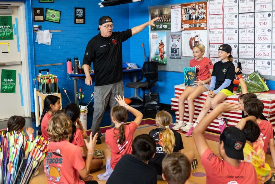 Coach Jim DeLine, gesturing with his left arm, chats with more than a dozen elementary school students inside the gym at Highland Park Elementary School.