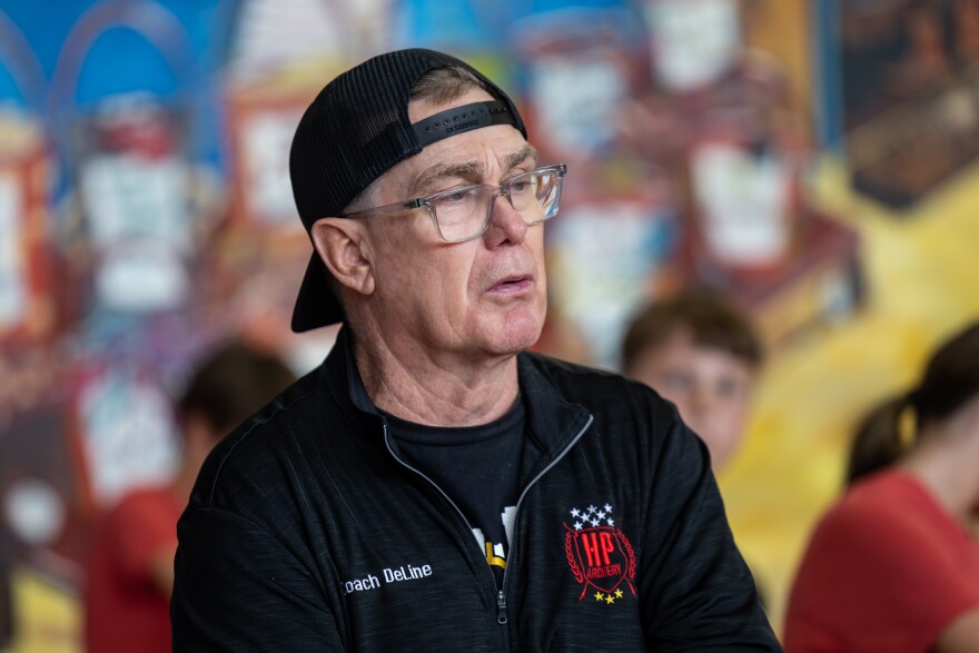 Coach Jim DeLine, wearing a backwards baseball hat and a sweatshirt that reads "Coach DeLine" leads his students through drills during after-school archery practice.