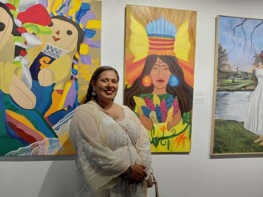 A woman wearing a white dress with gold trim stands in front of brightly colored Chicano art. Her hair is braided. 