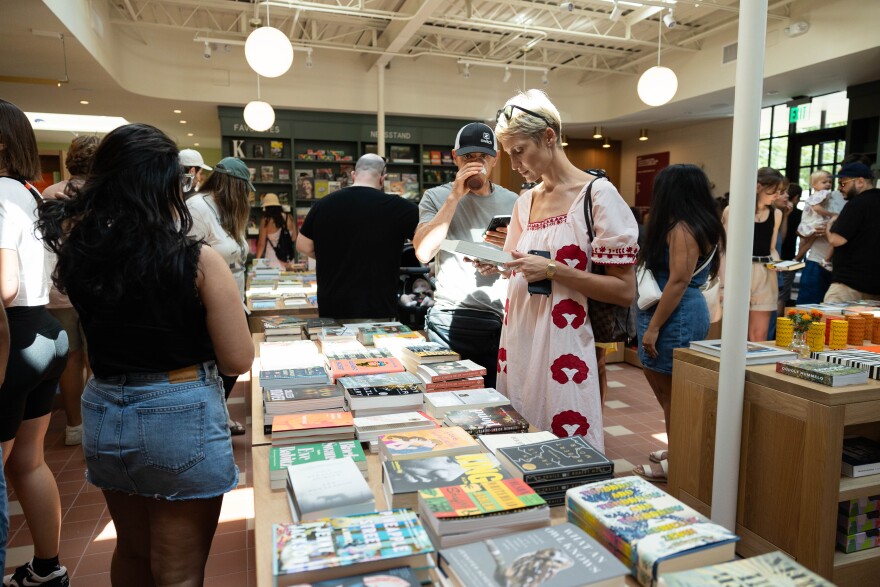 A woman reads the back cover of a book she is holding in her hands while standing in front of a table of other at a bookstore.