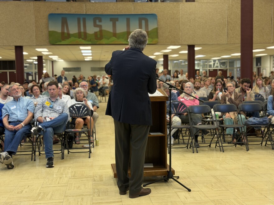 A man stands at a podium speaking to a crowd of people seated in a high school gym.
