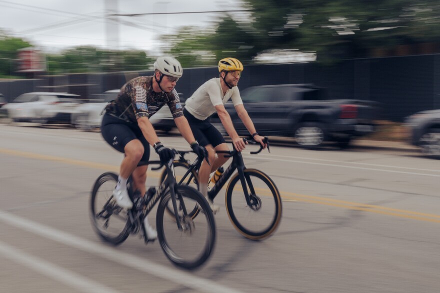 Two male cyclists are blurred while riding down a street.
