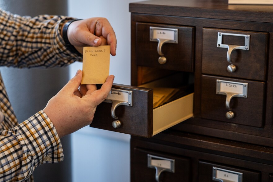 A person holds up a small yellow packet holding black bean seeds in front of an open drawer in a cabinet.