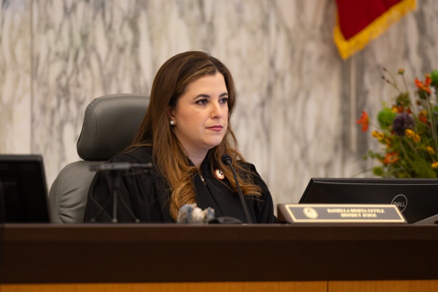 A woman in a judge's robe sites behind a desk and computer monitor. The desk has a plate with her name on it. Behind her is a marble wall.