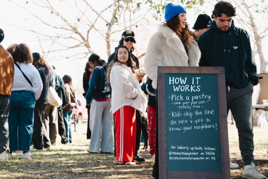 People wait in line in a park near a sign that list instructions on getting bakery goods.