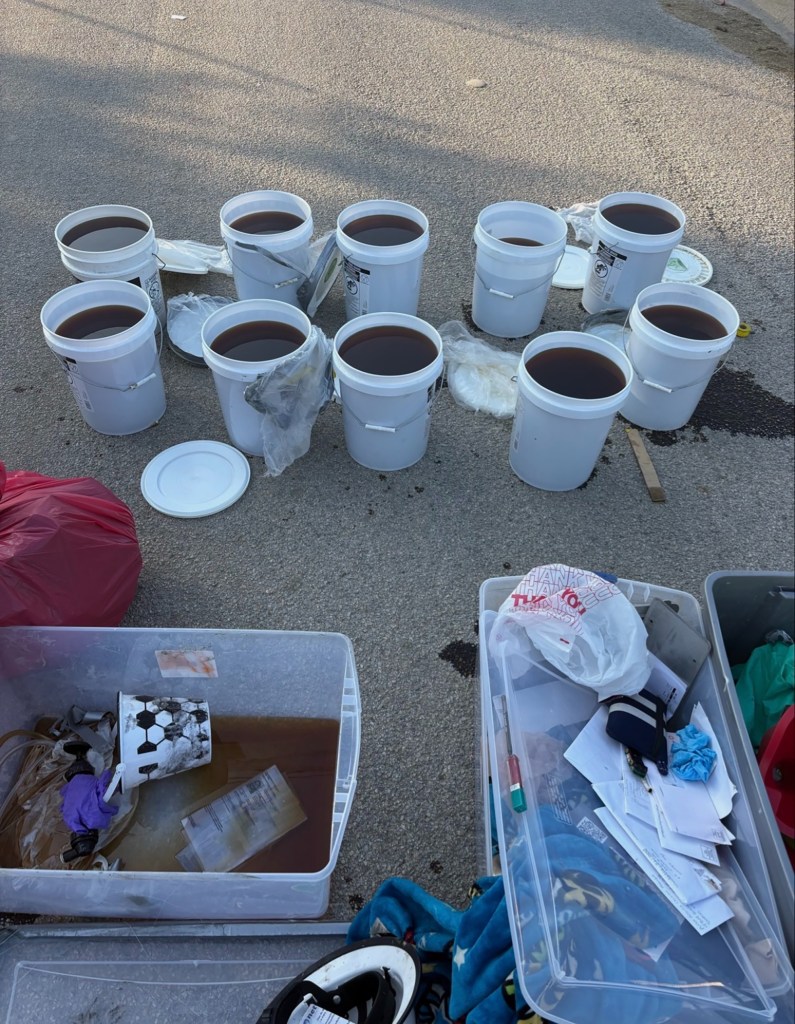 Ten white buckets containing liquid methamphetamine sit on the pavement, surrounded by other items removed from a car.