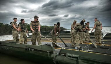 A dozen people in camouflage military uniforms and life jackets use ropes and other equipment to attach two sections of a floating bridge.