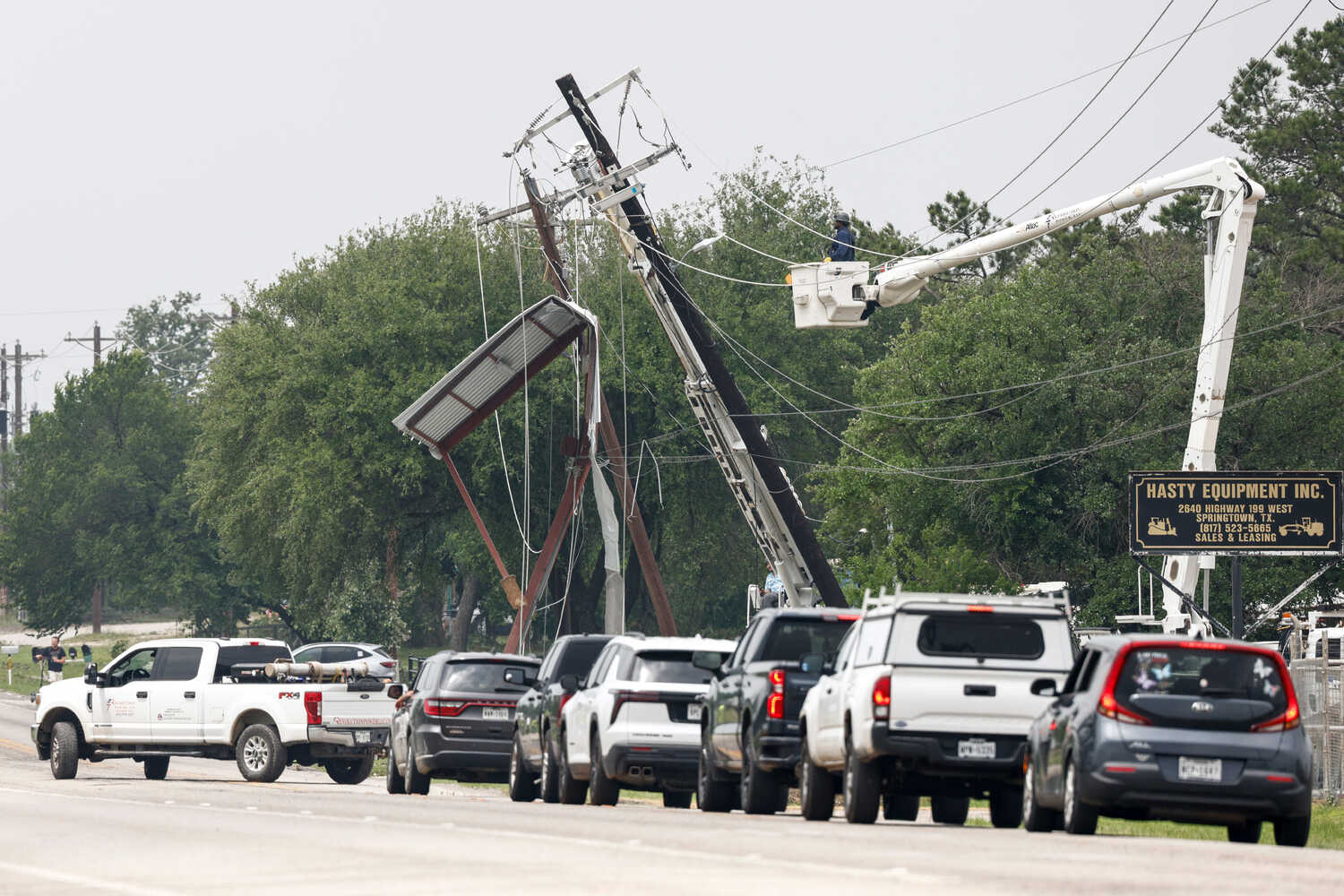 A utility works tends to a tilted and damaged telephone poll near a row of cars on a road. 