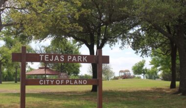 A wooden sign reading "Tejas Park" in front of several trees with a playground in distant background