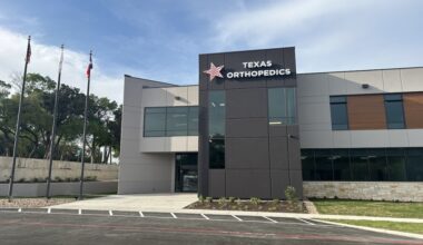 Texas Orthopedics logo with a red star displayed on a two story office building next to flag poles on a sunny day.