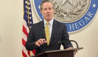 A man in a black suit and yellow tie speaks behind a wooden podium.