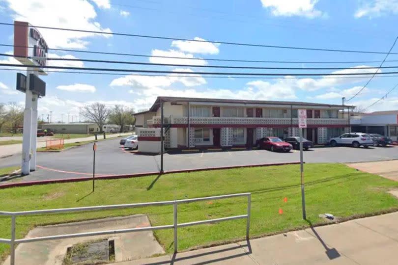 An image depicting an exterior view of a commercial establishment, featuring a grassy lawn, several parked vehicles in front, and a large signboard situated near the entrance. The scene is set under a blue sky with scattered clouds.