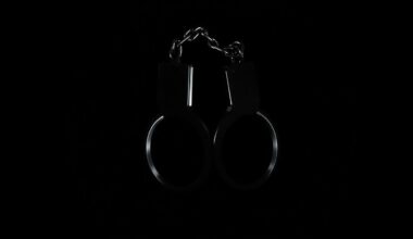 An extreme close-up photograph of a set of handcuffs against a pitch-black background, lit by a harsh, direct camera flash, conceptually representing the arrest of a local pastor on sexual assault charges.