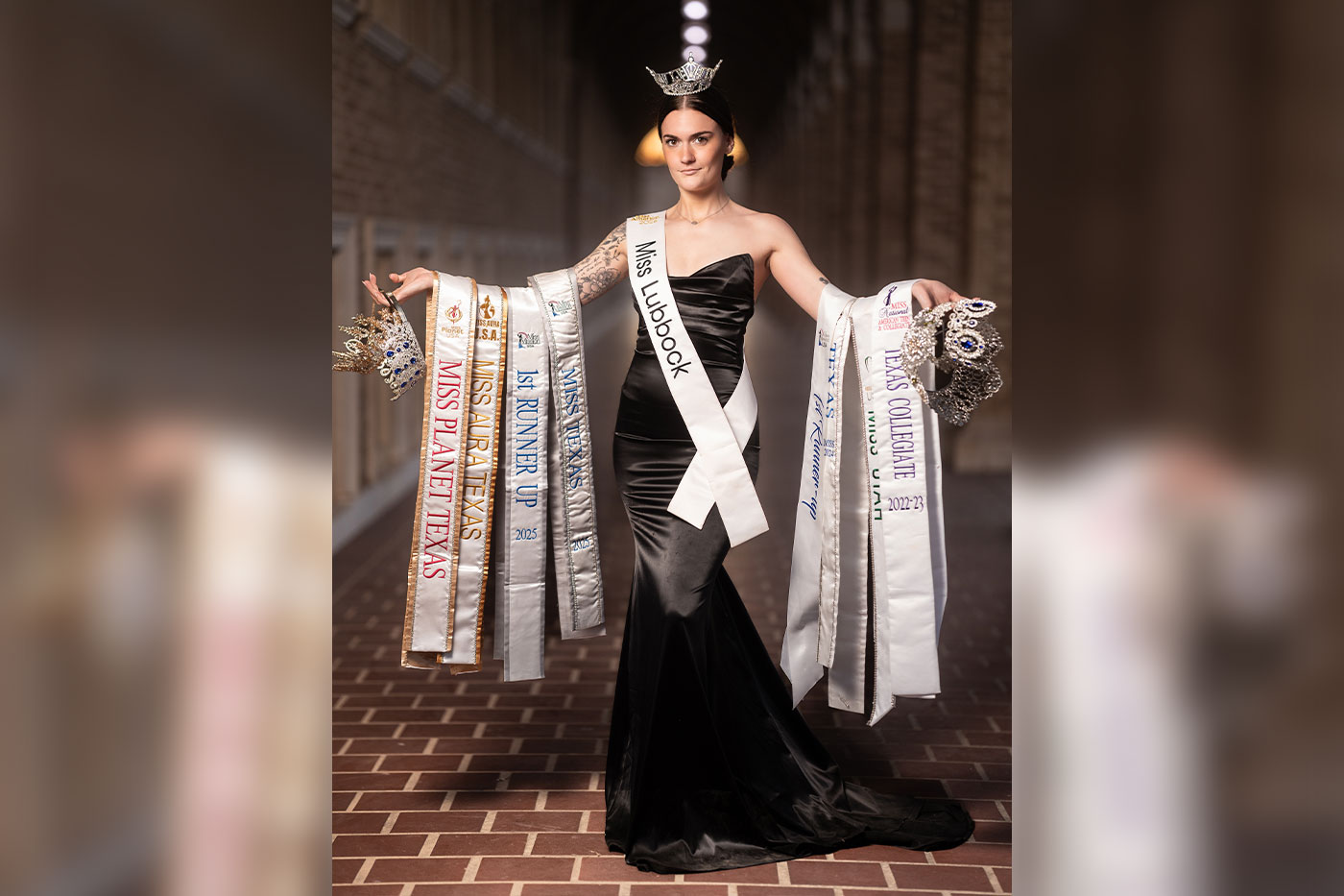 A crowned pageant contestant wearing a “Miss Lubbock” sash poses in a formal hallway, dressed in a black gown and displaying multiple competition sashes and tiaras in both hands.