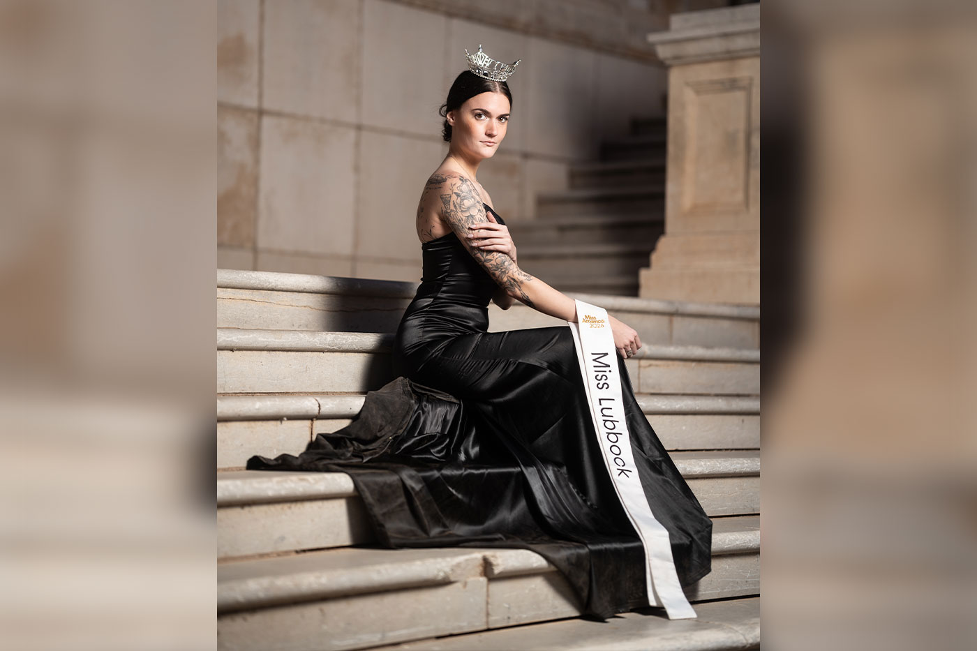 Miss Lubbock, wearing a crown and black gown and a tatoo sleeve on her right arm, poses seated on stone steps with her sash visible across her lap.