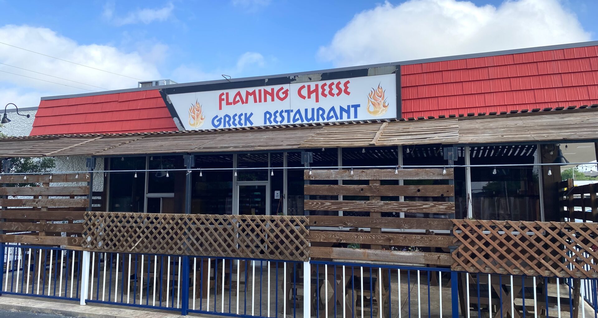 The exterior of Flaming Cheese, a Greek restaurant. There's a red awning and an enclosed patio.