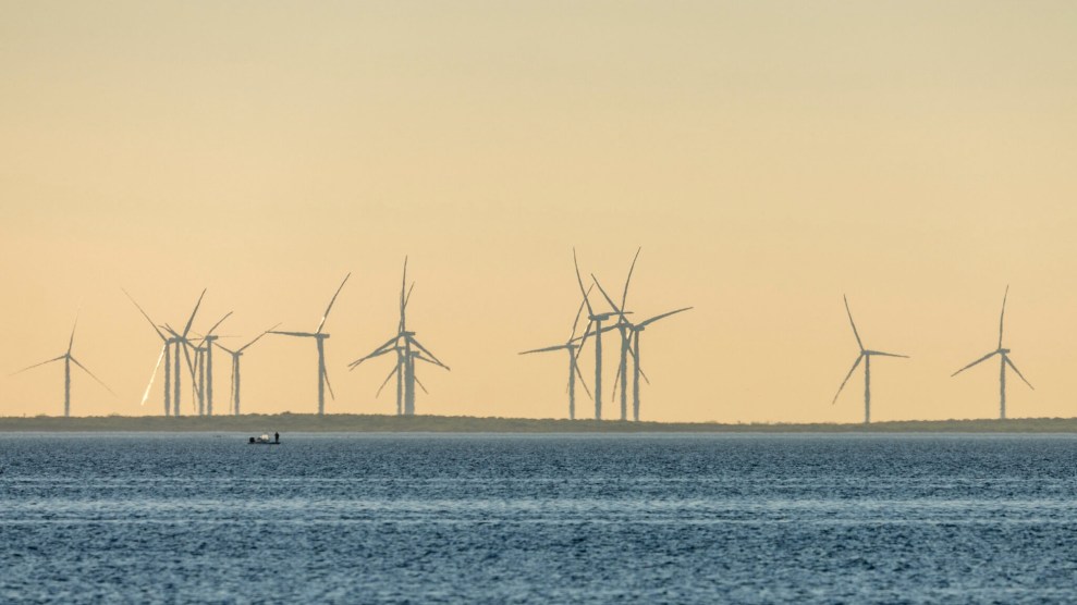 Wind turbines stand out of the water against on orange sky. A small fishing boat is far off in the water.