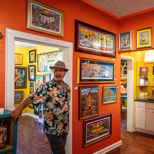 A photograph of artist and community advocate Hal Marcus standing inside a gallery with brightly painted walls, filled with paintings.