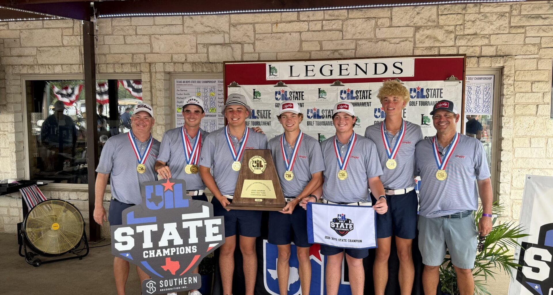 Seven young golfers in gray shirts with gold medals pose together in front of a Legends State Championships banner, holding a large trophy and banners.