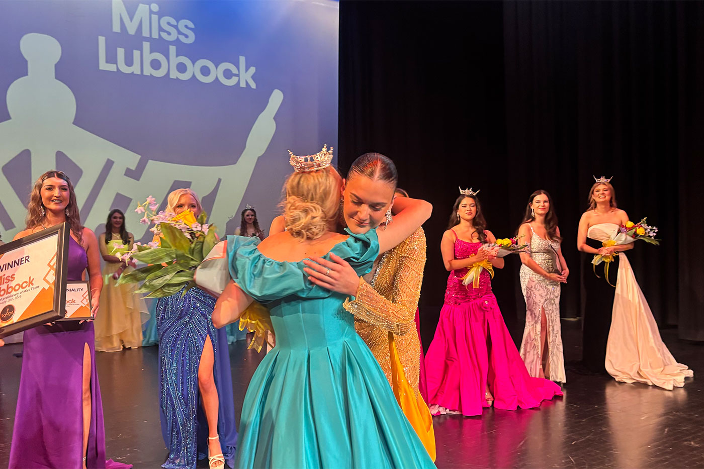 Miss Lubbock winner in a gold gown hugs a former titleholder during the crowning moment, with other contestants in evening gowns standing behind them.