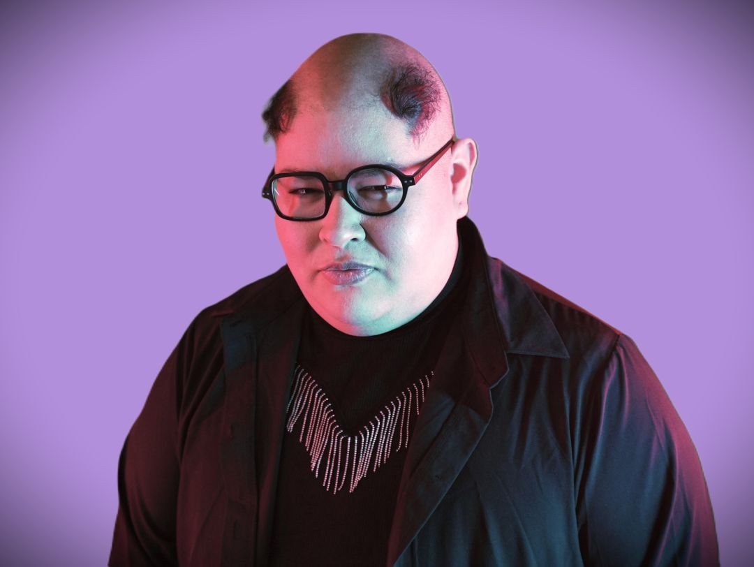 A person in a black shirt and glasses looks at the camera. There are purple lights shining on them.
