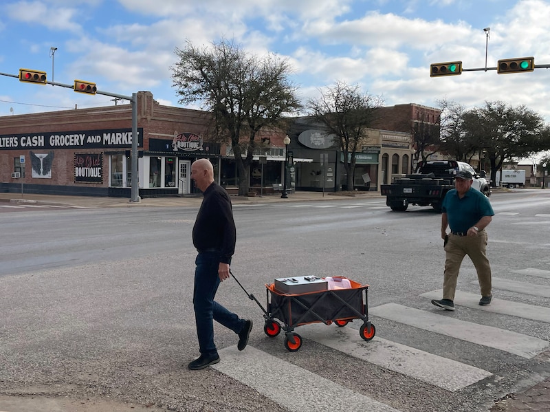 A photograph of two men walking down the street on a sunny day. One man is pulling a wagon with paper ballots inside.